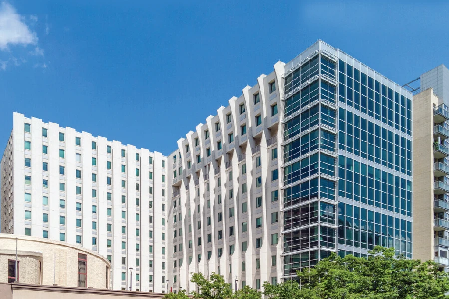 A modern multi-story building with a glass facade and concrete walls, set against a clear blue sky with some trees in the foreground.
