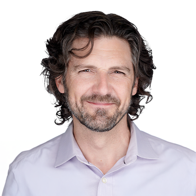 A man with wavy brown hair and a beard, wearing a light gray suit jacket and a light-colored shirt, poses against a white background.