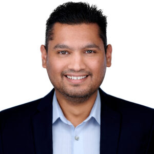 Smiling man with short dark hair and trimmed beard wearing a light blue collared shirt and a dark blazer, facing forward against a white background.